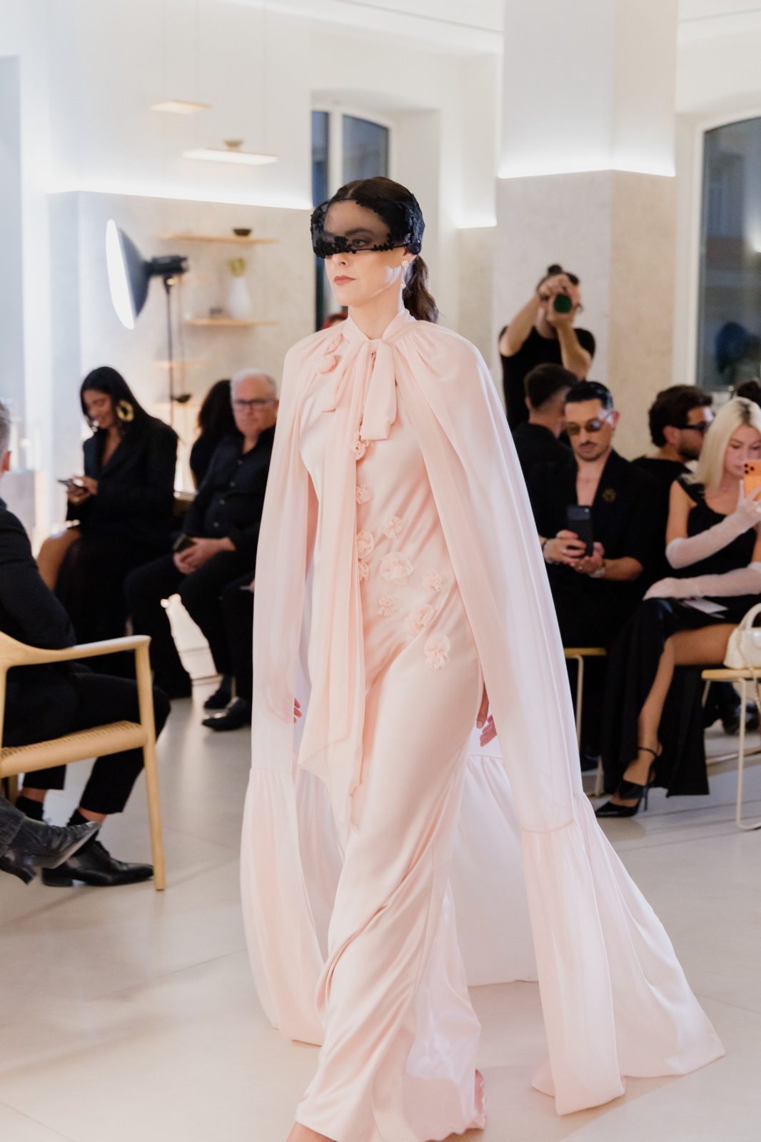 Modelo en pasarela con vestido rosa empolvado de línea entallada y capa larga con lazada al cuello.